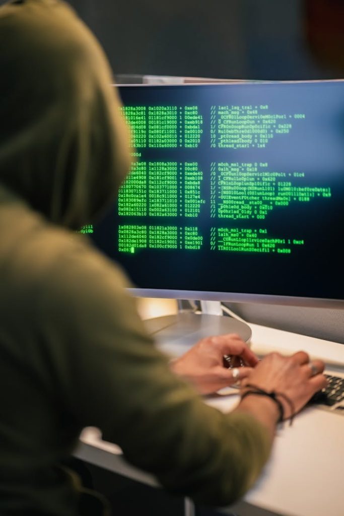 pexels photo 34258666 Person wearing a hoodie typing code on a computer screen with green text, depicting hacking scene.
